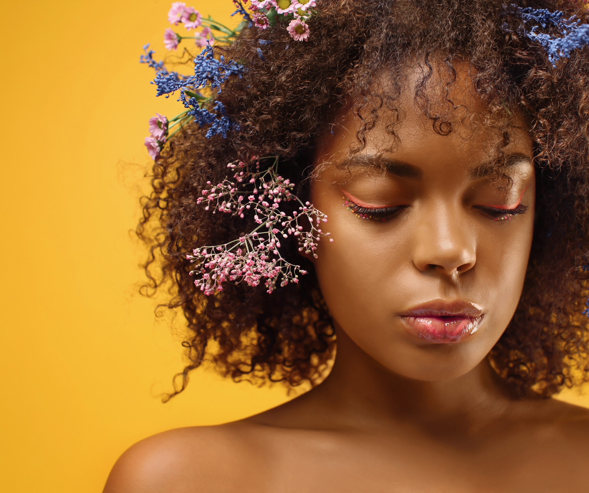 a black woman with pink, purple, and blue flowers tucked into her curls against a golden yellow background